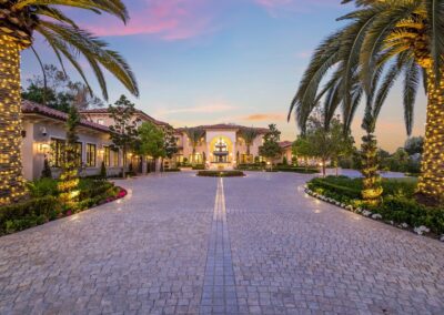 Luxurious mansion with a paved driveway lined by palm trees decorated with lights. The house is surrounded by greenery under a colorful evening sky.