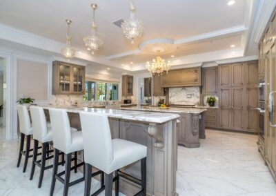 Luxurious kitchen with marble countertops, gray cabinetry, a large island with white barstools, and ornate chandeliers.